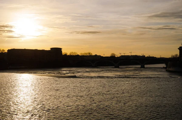 Yüzeyde dalgalar ve suda gün batımının yansımasının yer aldığı Garonne Nehri üzerindeki akşam manzarası Fransa 'nın Toulouse kentindeki Saint-Cyprien semtindeki binaların siluetleri