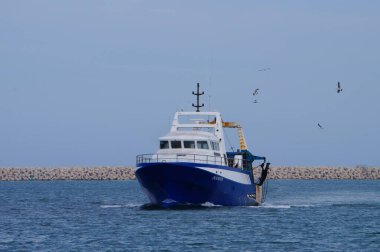 Port-la-Nouvelle, Fransa - 5 Nisan 2024 - Bir deniz gezisinden sonra modern bir balıkçı teknesi limana geri döner, ardından birkaç martı gelir; arka planda limanı çevreleyen taş iskele vardır.