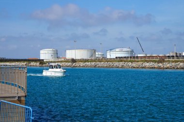 Port-la-Nouvelle, Fransa - 6 Nisan 2024 - Berre Nehri 'nin Akdeniz' e aktığı kanalda beyaz bir sürat teknesi, ticaret limanının petrol terminalinde dairesel tanklarla yüzüyor.
