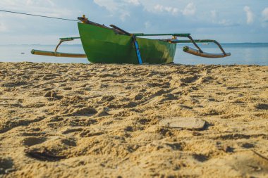 Yeşil ahşap bir tekne kumlu bir plajda oturur ve sakin dalgalar arka planda kıyıya vurur. Gökyüzü parlak..