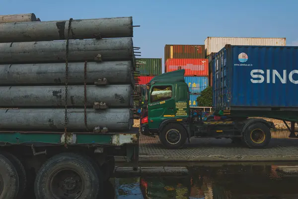 stock image Jakarta, Indonesia - May 9th, 2024. A large green truck with a yellow trailer loaded with concrete pipes is parked next to a blue container