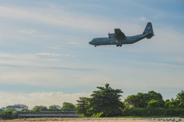 Balikpapan, Indonesia - June 14th, 2024. A  Lockheed C-130J Hercules military transport aircraft  is flying over a beautiful beach with lush green trees in the background. clipart