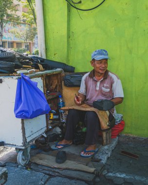 Balikpapan, Indonesia - June 22nd, 2024.  street vendor repairing shoes at an outdoor workshop. The image showcases the cobbler's tools, work in progress, and the bustling city life in the background. clipart