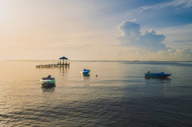 Sakin bir deniz manzarası ve sakin sulara demir atmış üç küçük tekne. Terası olan ahşap bir iskele yumuşak, bulutlu bir gökyüzünün altında denize doğru uzanır..