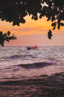 Gün batımında, gölgeli yapraklarla çerçevelenmiş, parlak, küçük bir tekne, yumuşak dalgalar üzerinde yüzer. Gökyüzünün sıcak ve canlı renkleri okyanusla harmanlanır..
