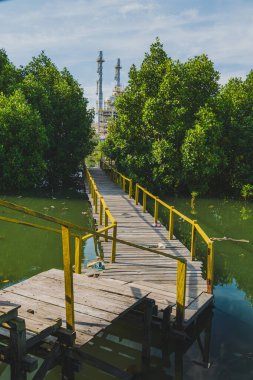 Balikpapan, Endonezya - 19 Nisan 2025. Petrol rafinerisinin kuleleri ve tesisleriyle mangrov bataklığını kesen ahşap bir patika..