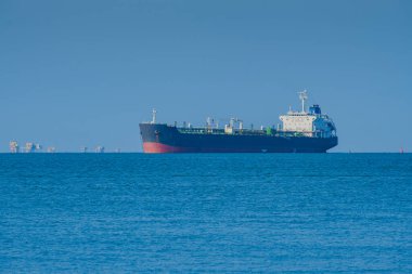 Siyah gövdesi ve kırmızı tabanı olan büyük, modern bir tanker gemisi, berrak bir gökyüzü altında engin, sakin bir mavi okyanusa yelken açar. Uzakta, Balikpapan, Endonezya 'da çeşitli kıyı petrol platformları görülebiliyor..
