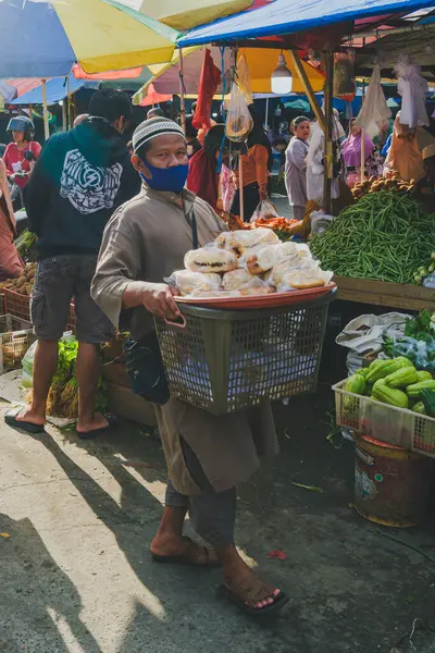 Balikpapan, Endonezya - 19 Nisan 2025. Bir sokak satıcısı Pandan Sari geleneksel pazarında taze sebzeler ve renkli şemsiyelerle çevrili bir sepet paket ekmek taşıyor..