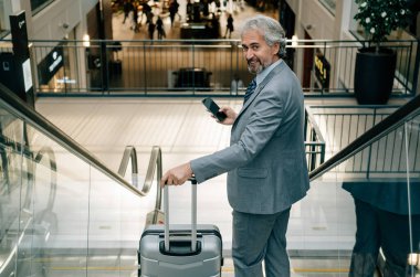 Zarif gri takım elbiseli mutlu bir iş adamı telefonuna mesaj yazıyor ve bavuluyla yürüyen merdivenden aşağı inerken kameraya bakıyor.