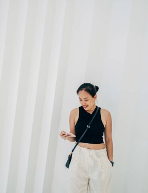 Beyaz geometrik duvarın önünde duran mutlu kadın cep telefonuyla bir şeyler okuyor ya da izliyor.