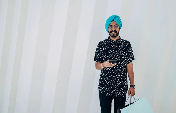 Portrait Of Happy Indian Man Using A Mobile Phone While Holding Shopping Bag And Standing In Front Of White Geometric Wall