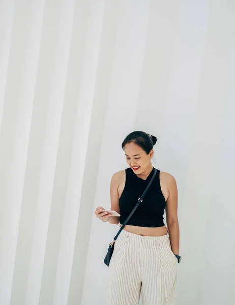 一个快乐的女人站在白色的几何图形墙前 一边看一边看手机上的东西 — 图库照片