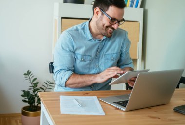 Ofis masasında bilgisayar, kağıt ve kalemle çalışırken gülümseyen işadamı tabletine yazı yazıyor.