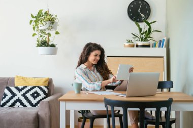 Gülümseyen iş kadını bir fincan kahve, dizüstü bilgisayar, kağıt ve kalemle ofis masasında çalışırken tabletinde rapor okuyor.