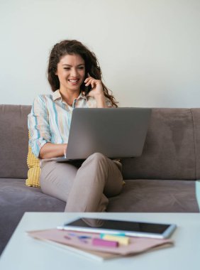 Mutlu iş kadını cep telefonuyla konuşuyor ve oturma odasındaki kanepede otururken dizüstü bilgisayarda iş raporu yazıyor.