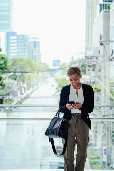 Happy business woman typing text message on her mobile phone while standing on a modern office terrace with city view