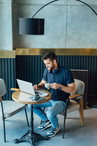ノートパソコンのキーボードでビジネスレポートを入力し コーヒーショップに座っている間にエスプレッソコーヒーのカップを飲む幸せな男の角度のビュー ストックフォト