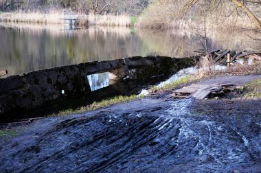Batan beton duvarlı göl. Dar lastik izleriyle çamurlu zemin. Ahşap palet köprüsünden geçen küçük bir dere. Uzak kıyılardaki kuru sazlıklar, köprünün ötesindeki patika devam ediyor.