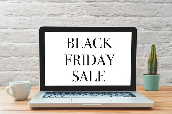 Black friday sale is written on the screen of a laptop. The computer is placed on a wooden desk with an espresso cup and a cactus in the background. The notebook is open.
