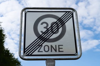 Bu trafik levhası 30 km / s 'lik alanın sonu olduğunu gösteriyor. Bir direğin üzerindeki beyaz yol tabelasının gri 30 ve siyah çizgileri var. Arka planda gökyüzü ve bulutlar görülebilir.. 