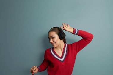 Güzel kız en sevdiği şarkıyı söylüyor, kablosuz kulaklıkla müzik dinliyor, gülümsüyor ve dans ediyor, mavi arka planda duruyor.