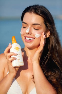 Yüzüne güneş kremi süren bir kadın gülümsemesi. Cilt bakımı. Vücut Güneşi koruması. Güneş kremi. Ciltte kadın yağı nemlendirici losyon.