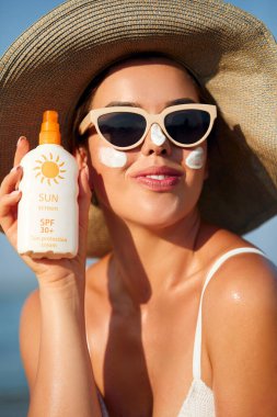 Yüzünde güneş kremi olan güzel genç bir kadın. Sahilde güneş kremi şişesi tutan bir kız. Kadın şapka cilt üzerinde nemlendirici losyon uygulayarak. Cilt bakımı. Güneş koruması. Güneş