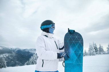 Kar sörfçüsü beyaz kış kıyafetleri içinde kar kaykayı ile karlı bir dağda manzaralı bir kış sporu macerasına hazır.