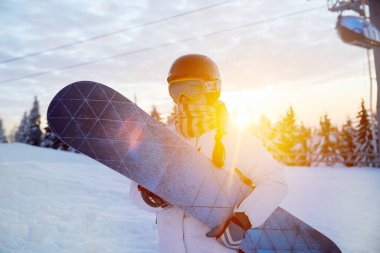 Dağların ve Günbatımının Arkasındaki Kadın Portresi. Snowboard tutuyor ve kayak gözlüğü takıyor.
