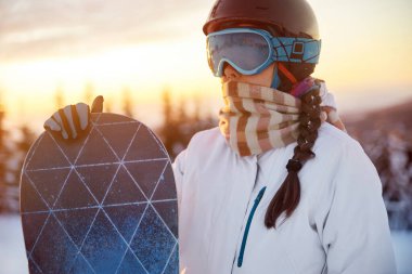 Dağların ve Günbatımının Arkasındaki Kadın Portresi. Snowboard tutuyor ve kayak gözlüğü takıyor. Yansımalı Snowboardcunun Kayak Gözlüğü.