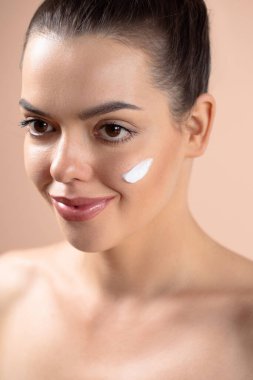 Güzel bir kadın bej arka planda parmakları yanağında beyaz krem sürüyor. Doğal çıplak makyajlı güzellik modeli portresi. Spa, cilt bakımı, sağlık. Kapat..