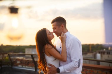 Gün batımında çatıda iki kişi öpüşür. Sıcak güneş ışığı, kucaklaşırken yüzlerinde altın bir parıltı salıyor, şehrin ufuk çizgisi ile çevrili..