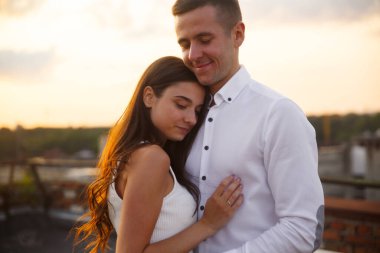 Bir erkek ve kadın arka planda güneş batarken çatıda hassas bir anı paylaşıyorlar. Sıcak ifadeleri, canlı bir şehir ortamında aşkı ve samimiyeti ifade eder..