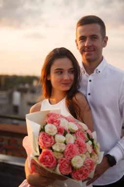 Gün batımında panoramik şehir manzarasıyla çevrili bir çift çatıda yan yana durur. Kadın canlı bir buket pembe ve beyaz çiçek tutuyor. Sevgi ve neşe taşıyor..