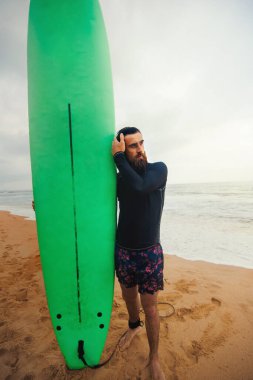 Plajın yanında sörf tahtası olan genç sakallı bir adam. Yaz günü sörf tahtası olan bir adam..