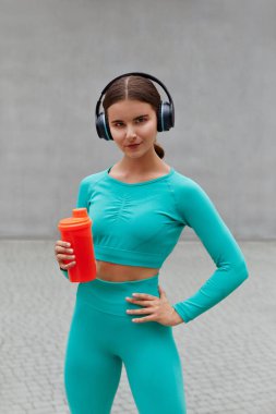 Beautiful female runner standing outdoors holding water bottle and using wireless headphones. Fitness woman taking a break after running workout