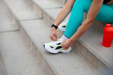 Açık hava ortamında, canlı spor kıyafetleri içinde bir birey somut basamaklarda oturur, egzersiz oturumuna başlamadan önce ayakkabı bağcıklarını düzeltir. Yakında bir su şişesi var..