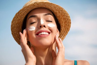 Kadın gülüşü yüzüne nemlendirici güneş kremi sürüyor. Cilt bakımı. Vücut Güneşi koruması. Güneş kremi. Şapka lekeli bir kadın cilde nemlendirici losyon sürüyor. Plajda elinde güneş kremi şişesi tutan bir kız. Güneş yanığı