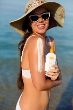 Omzuna güneş kremi süren genç ve güzel bir kadın. Kız şık bir güneş gözlüğü takıyor ve sıcak bir yaz gününde güneşin tadını çıkarıyor.. 