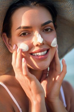 Yüzüne güneş kremi süren bir kadın gülümsemesi. Cilt bakımı. Vücut Güneşi koruması. Güneş kremi. Şapkalı bir kadın cilde nemlendirici losyon sürüyor.