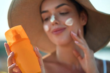 Yüzünde güneş kremi olan güzel genç bir kadın. Plajda elinde güneş kremi şişesi tutan ve cildine nemlendirici losyon süren bir kız. Güneş koruması. Güneş yanığı.
