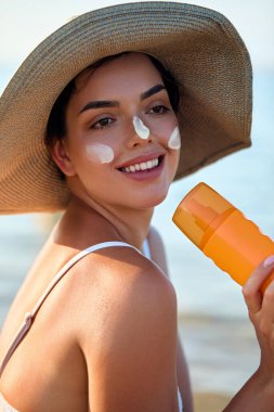 Yüzünde güneş kremi olan güzel genç bir kadın. Plajda elinde güneş kremi şişesi tutan ve cildine nemlendirici losyon süren bir kız. Güneş koruması. Güneş yanığı.