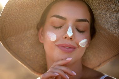Yüzünde güneş kremi olan güzel genç bir kadın. Şapkalı bir kadın cildine güneş kremi sürüyor. Nemlendirici losyon sürüyor. Cilt bakımı. Güneş koruması. Güneş yanığı