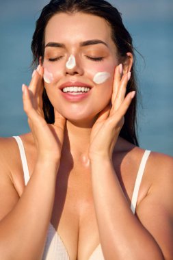 Güzel bir kadın bronzlaşmış bir yüze krem sürüyor. Deri ve vücut bakımı kavramı... Kız cilt için güneş kremi kullanıyor. Güneş kremi ya da nemlendirici güneş kremi tutan bir kadının portresi.