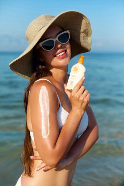 Bikinili kız cildine güneş kremi sürüyor. Şapka lekeli nemlendirici losyonlu bir kadın. Sahilde elinde güneş kremi şişesiyle gülümseyen güzel bir kadın. Deri ve Vücut Bakımı Konsepti. Güneş koruma kavramı. 