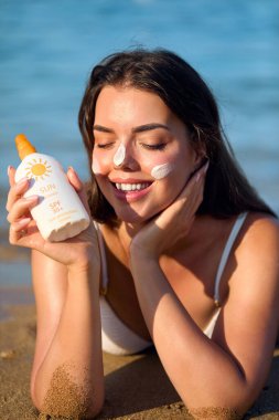 Gülümseyen kadın plajda dinlenirken yüzüne güneş kremi sürüyor, güneş korumasının keyfini çıkarıyor ve sağlıklı bir yaz cilt bakımını teşvik ediyor. Deri Bakımı Konsepti