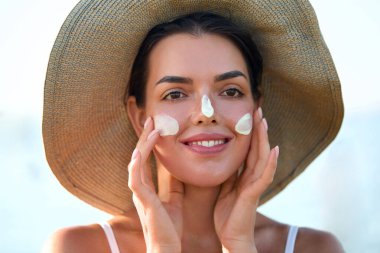 Genç ve güzel bir kadın güneş kremi sürüyor ve kendi yüzüne dokunuyor. Cilt bakımı. Güzellik Konsepti. Şapkalı bir kadın cildine güneş kremi sürüyor. Cilt Koruma ve Dermatoloji