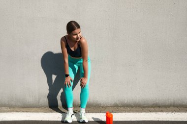 Sporcu bir kadın antrenmandan sonra mola veriyor, duvara yaslanıyor, spor kıyafetlerini giyiyor, su şişesiyle su içmeye odaklanıyor.