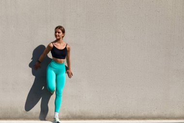 Aktif bir kadın basit bir duvara yaslanıyor, modaya uygun spor kıyafetleri giyiyor, canlı bir spor ve sağlıklı yaşam tarzını somutlaştırıyor.