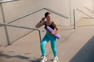 Dışarıdaki merdivenlere tırmanırken yoga minderi taşıyan genç atletik bir kadın, canlı aktif giysiler içinde, spor yaşam tarzını ve modern şehir ortamında sağlık ve sağlığa olan bağlılığını sergiliyor..